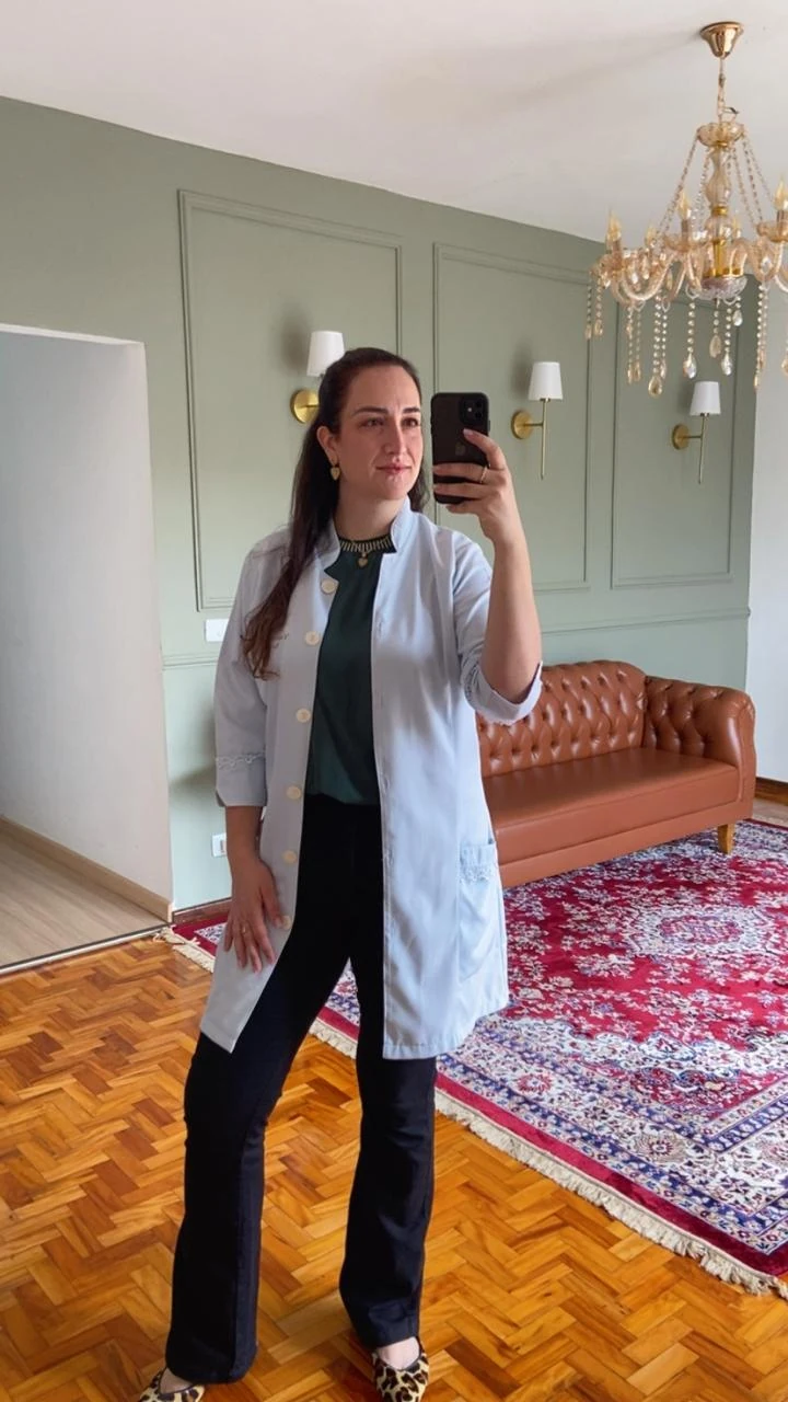 Vanessa Maichaki vestindo jaleco branco em uma sala elegante, diante de uma parede verde e sofá caramelo